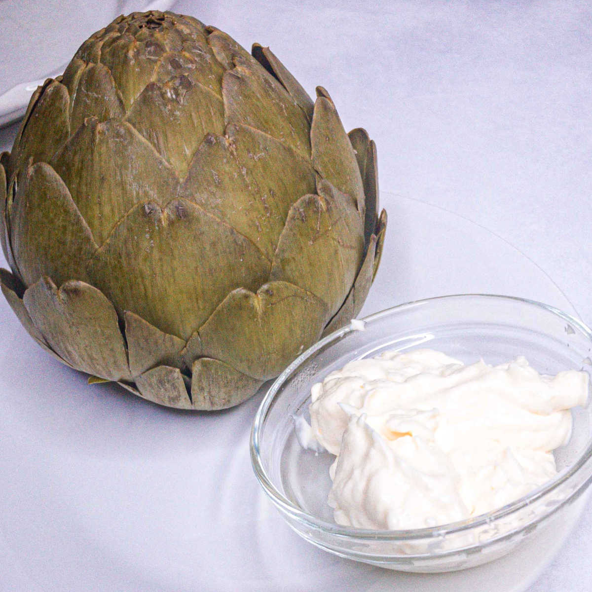 Cooked artichoke on a white plate with a small bowl of mayonnaise dip.