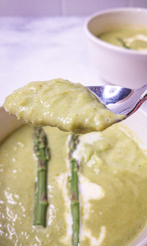 Closeup of a spoon loaded with creamy asparagus soup with bowls of soup in the background.