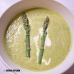 top down view of a bowl of creamy asparagus soup with a drizzle of cream in it as well as two tips of asparagus stalks.
