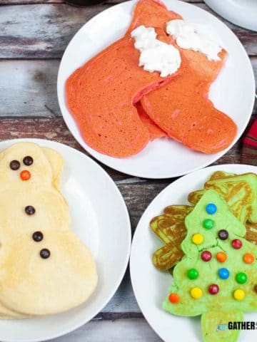 Tops down view of a plate of red stocking pancakes, plate of snowmen pancakes, and a plate Christmas tree pancakes.