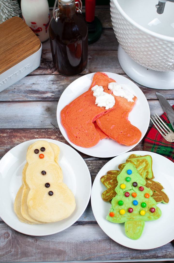 Three plates, one with red stocking pancakes, one with green Christmas tree pancakes, and one with white snowmen pancakes.