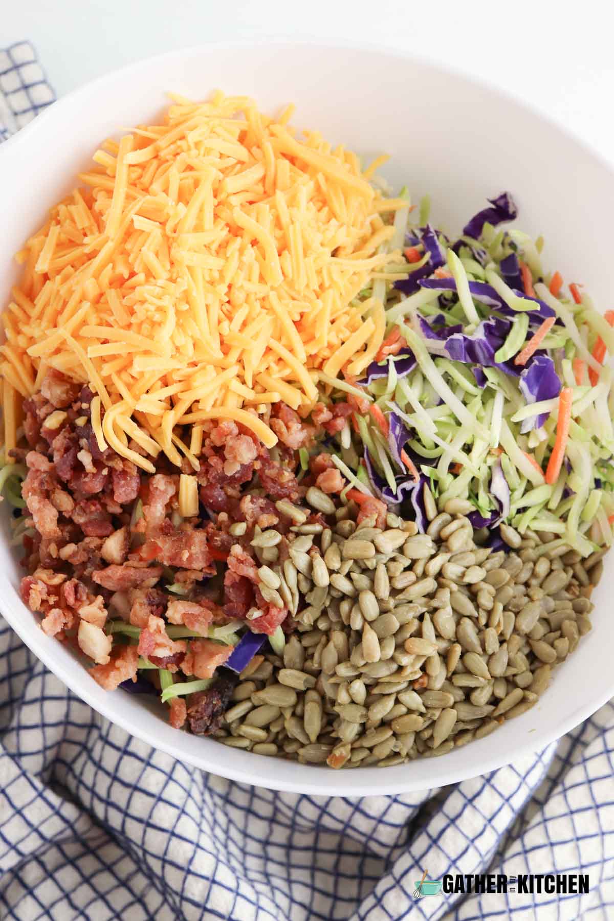 Ingredients for broccoli slaw in a bowl: shredded cheese, broccoli slaw, sunflower kernels, and crumbled bacon.