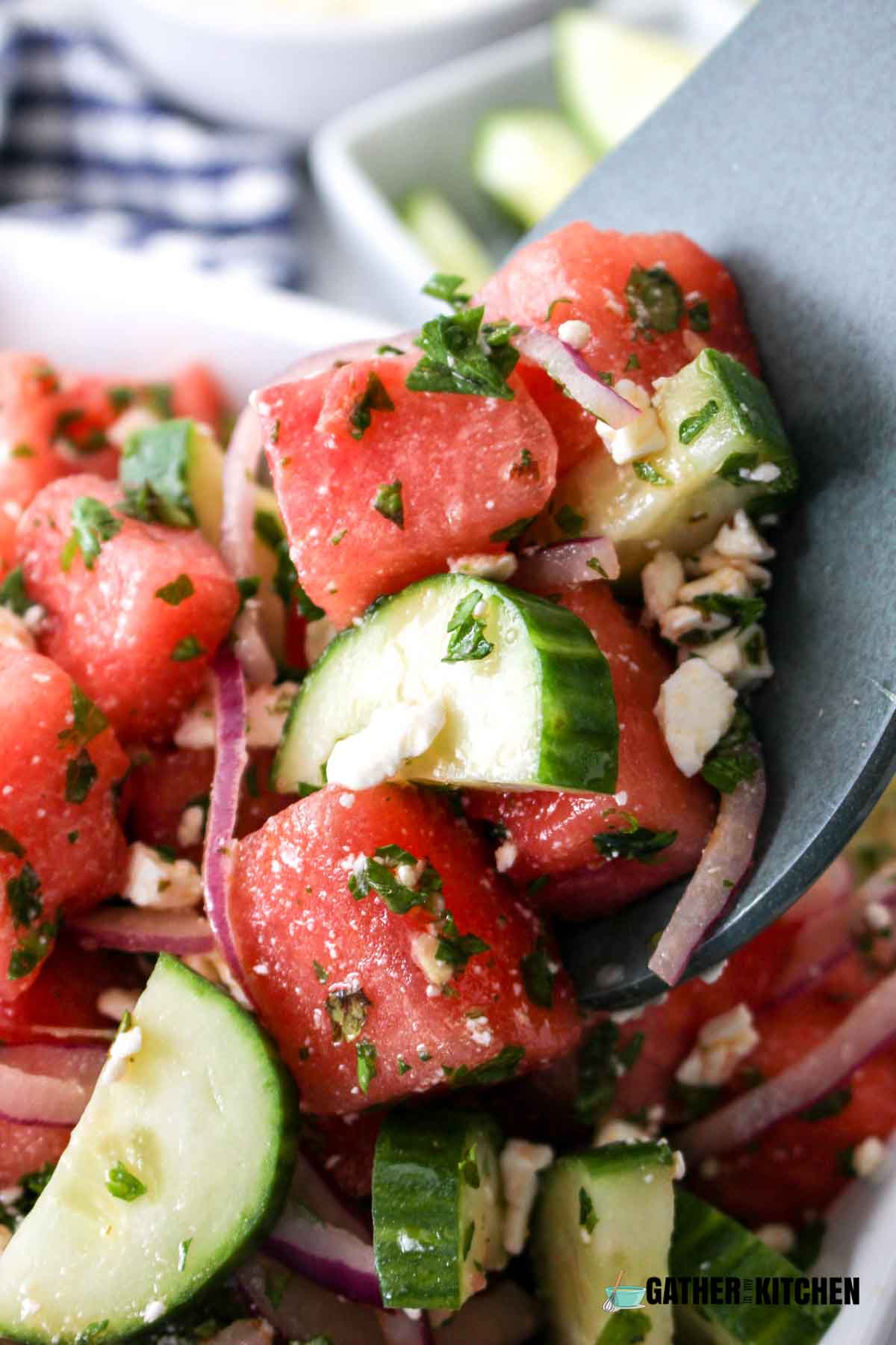 Closeup of a spoon holding watermelon salad - you can see watermelon, pieces of red onion, chopped cucumbers with herbs and feta sprinkled throughout.