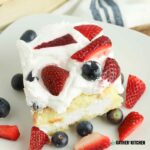 Angled shot of a slice of twinkie shortcake on a plate with strawberries and blueberries on top as well as around the slice.