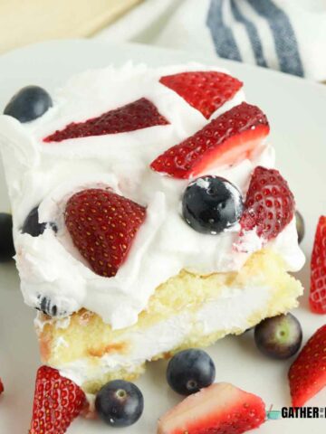Angled shot of a slice of twinkie shortcake on a plate with strawberries and blueberries on top as well as around the slice.