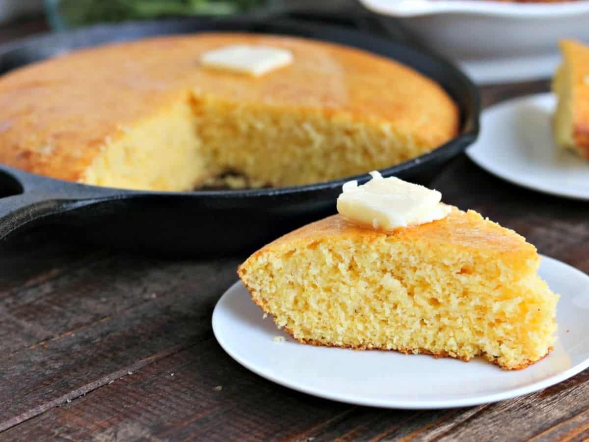 Piece of corn bread on a plate with the rest of the corn bread in a skillet behind.