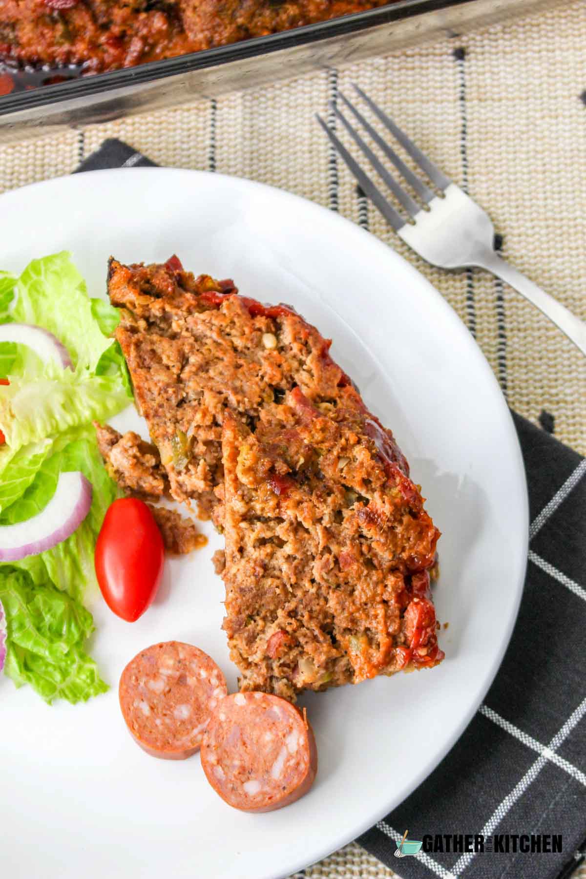 A slice of cajun meatloaf on a plate with salad and sausages.