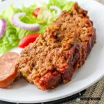 Closeup of a slice of cajun meatloaf on a plate with a salad and sausage on the side.