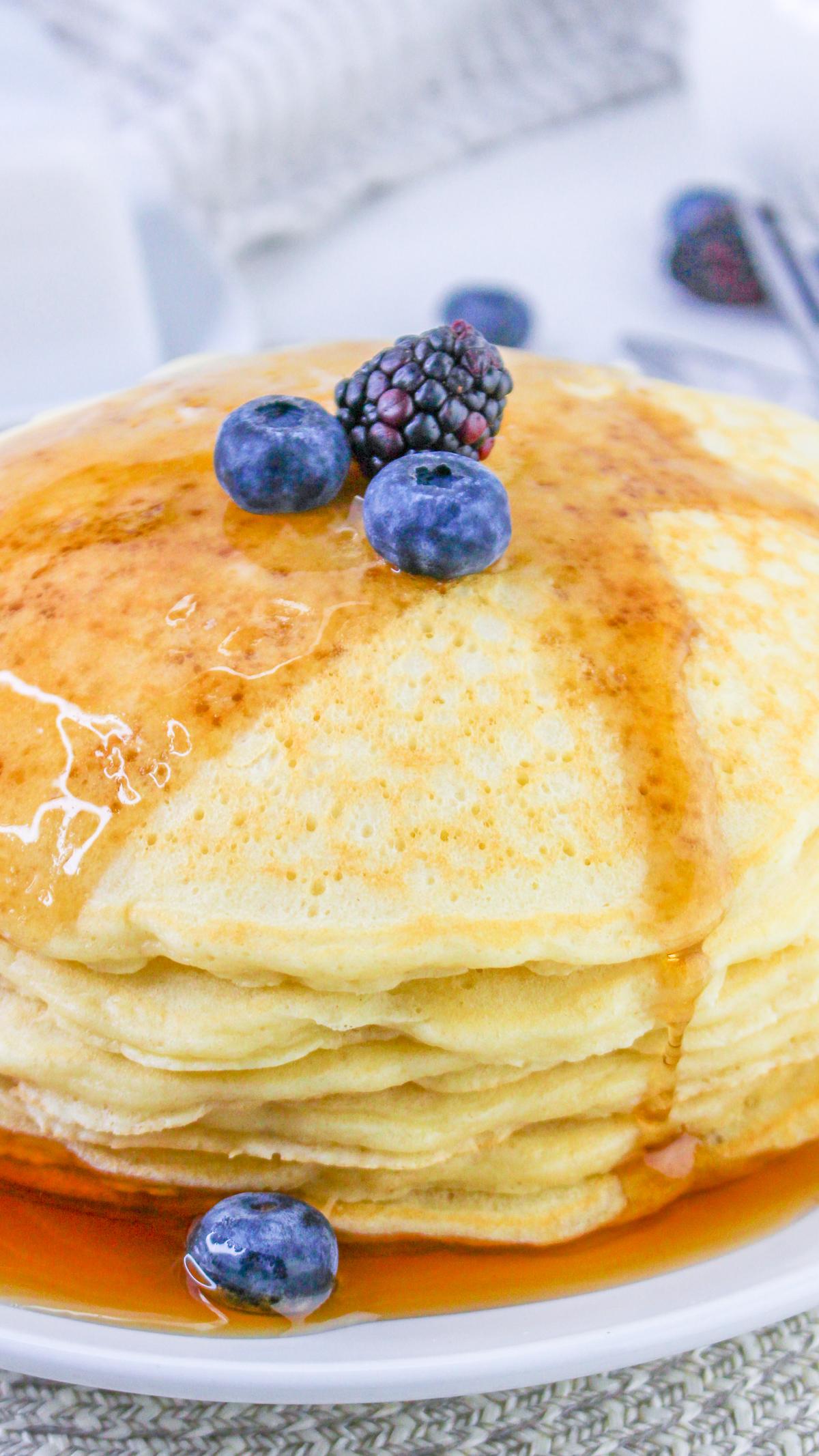 Stack of sweet cream pancakes on a plate with a few berries on top and covered in syrup.