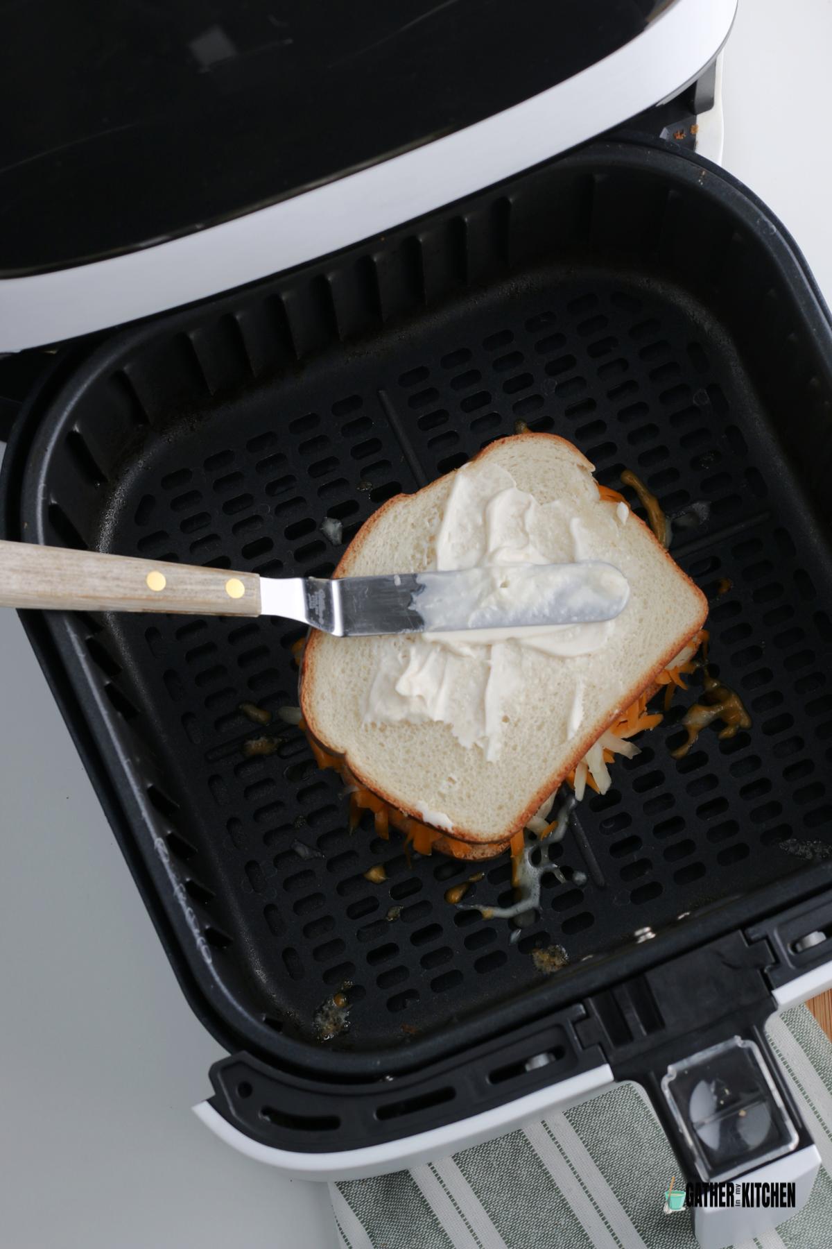 Uncooked sandwich in air fryer basket and spreading mayo over the top slice of bread.