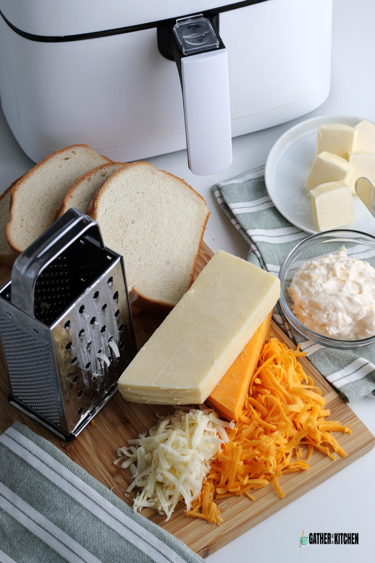 Air fryer, plate of butter, small bowl of mayonnaise, cheddar and monterey Jack cheese in block form plus a bit of shredded, cheese grater, and slices of sour dough bread.