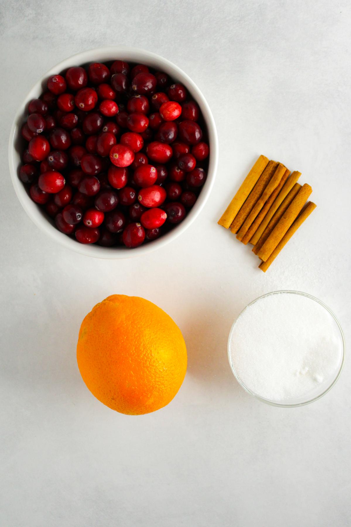Ingredients for cranberry sauce: fresh cranberries in a bowl, cinnamon sticks, small bowl of white sugar and an orange.