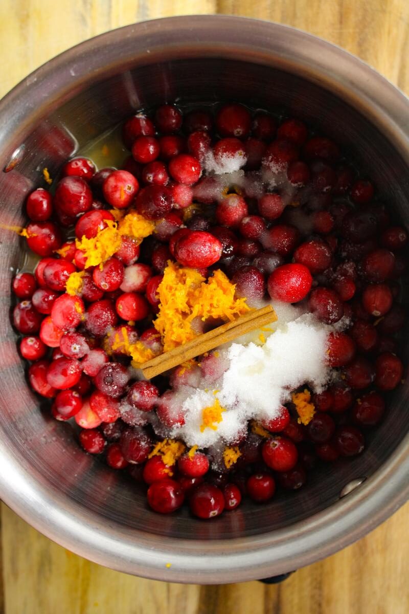 Top down view of a sauce pan with orange juice, fresh cranberries, orange zest, white sugar, and a cinnamon stick in it.